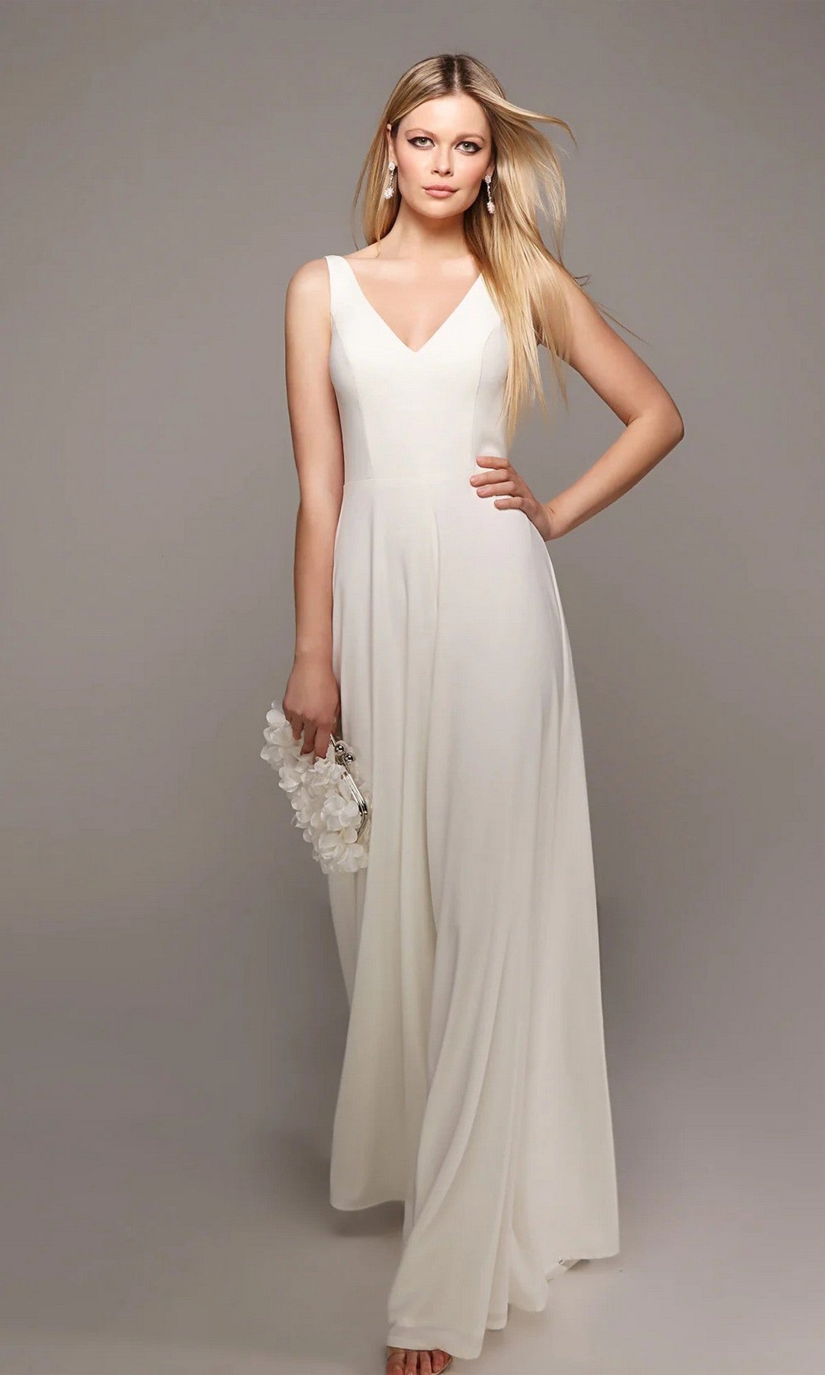 A woman in the Alyce 70021 long white jumpsuit with wide-leg pants holds a white floral clutch, posing against a plain gray background.